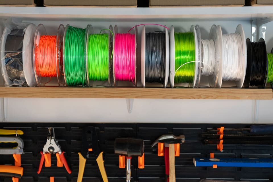 A shelf of 3D printing filament spools in orange, greens, hot pink, black and white above a pegboard of tools