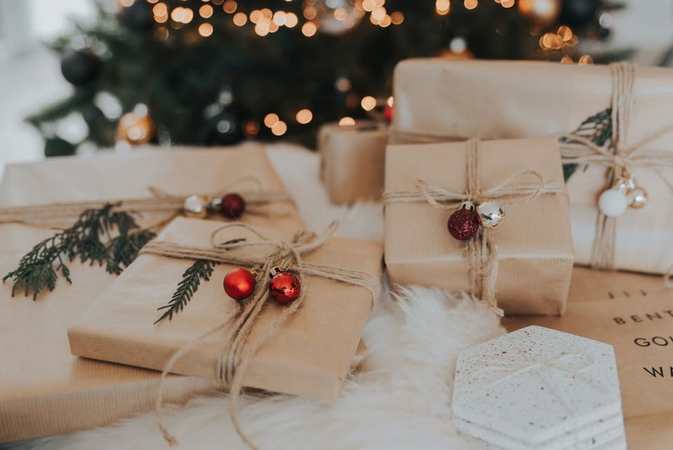 Brown-paper wrapped presents tied with twine, red mini baubles and fir sprigs, on a sheepskin under a lit Christmas tree