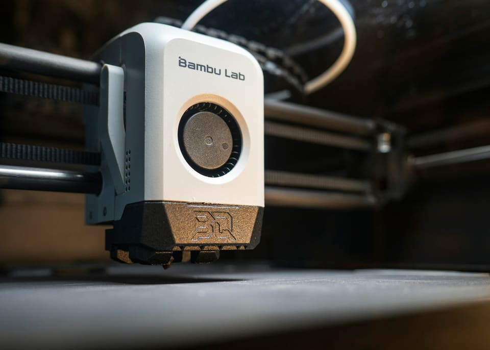 Close-up of a white Bambu Lab 3D printer toolhead moving over a dark build plate inside an enclosed printer