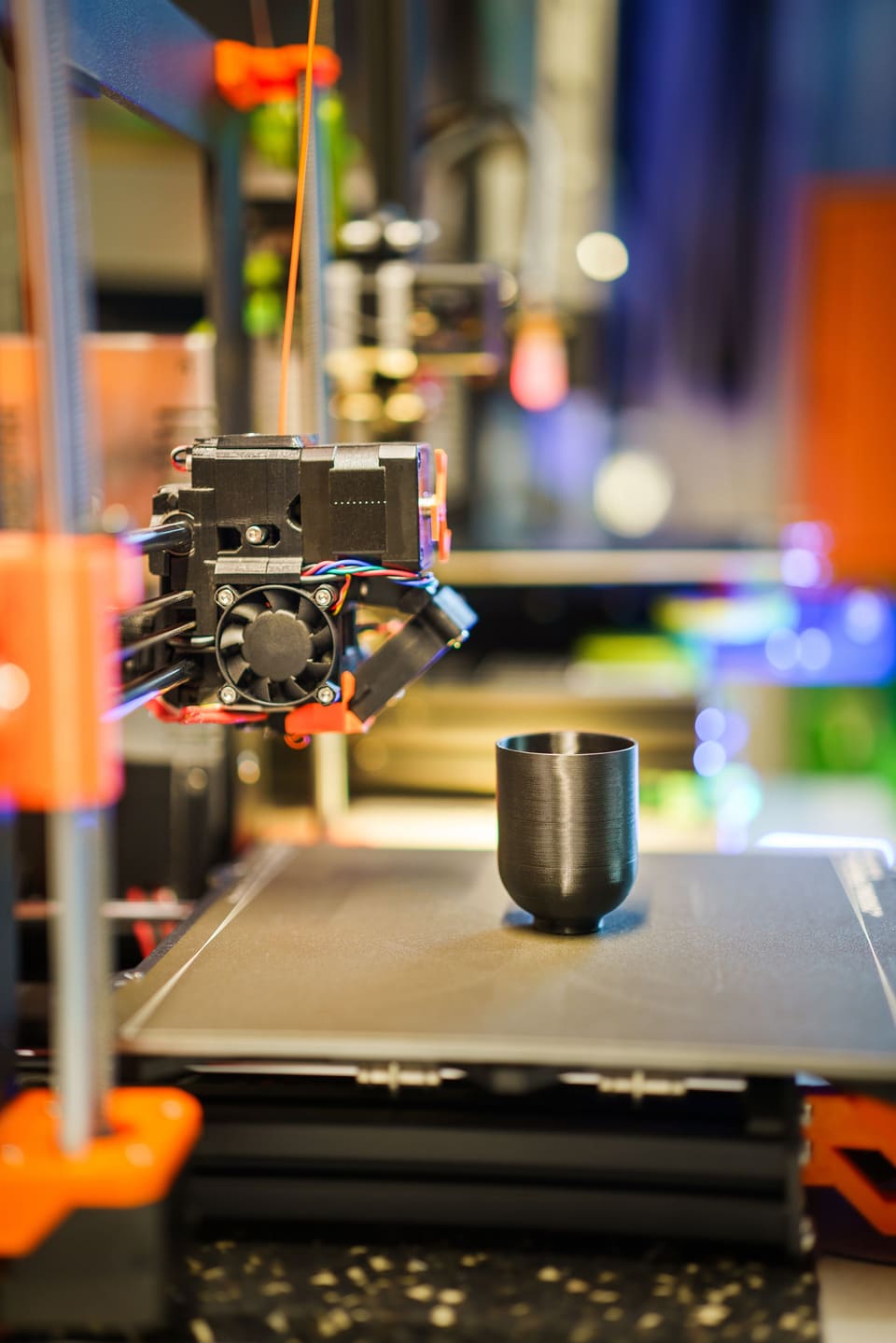 A just-finished black printed cup standing on a 3D printer bed beside the print head, workshop lights blurred behind